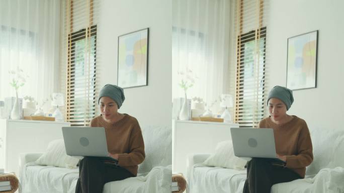 一名年轻的亚洲癌症患者，戴着头巾，坐在一间光线充足、植物装饰的房间里的沙发上，舒舒服服地摆弄着笔记本