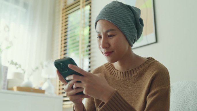 一个患癌症的年轻亚洲女人的特写，她穿着便服，戴着头巾，坐在舒适的沙发上玩手机，周围是一个明亮的，充满