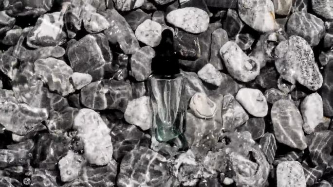 带移液管的玻璃化妆品瓶躺在海滩上的鹅卵石上。海浪拍打着鹅卵石。水面纹理。太阳和阴影。女性化妆品护肤品