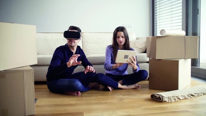 一对年轻夫妇带着平板电脑和VR眼镜坐在地板上，搬进了新家。