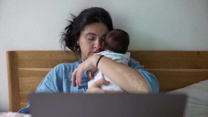 疲惫的母亲肩上抱着熟睡的新生儿，坐在床上拿着笔记本电脑，描绘着在日常生活中平衡工作和育儿的挑战和美好
