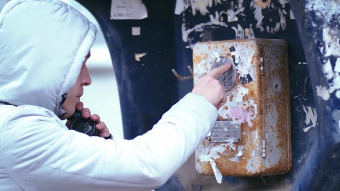 街头呼叫:在生锈的城市角落，傍晚阴影，犯罪现场，戴兜帽的人使用旧付费电话