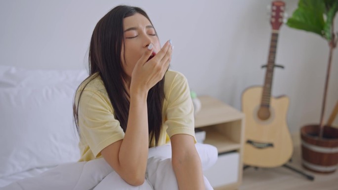 亚洲年轻困倦的女子在清晨从床上起床。疲惫迷人的女孩穿着睡衣感到心烦意乱，疲惫不堪，不想在家里的卧室里