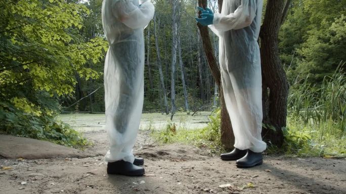 穿着白色防护服的人们讨论要检测的水样。医务人员在河岸附近工作，进行分析。化学危险，自然研究。