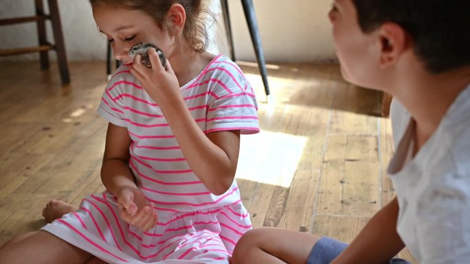 小女孩愉快地抱着一只小鸟，男孩在一旁观看，在舒适的室内环境中创造了童年好奇心和与动物联系的温馨时刻。