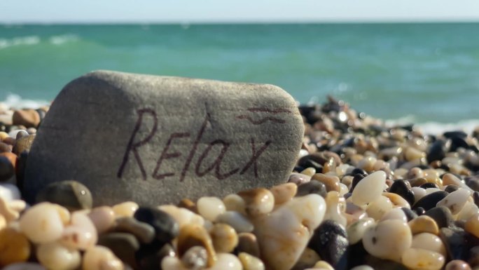 字放松手写在石头上的夏天卵石滩溅起的海浪。夏季海滩度假背景。