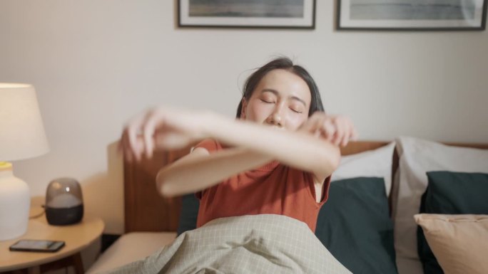 亚洲女性在床上醒来，用AI智能手机应用程序在日出时打开智能家居的窗帘