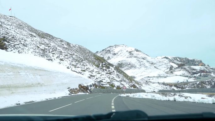 驱车穿过奥地利大格洛克纳地区积雪覆盖的高山地带，沿途蜿蜒的道路贯穿群山，展现了高海拔地区崎岖的美景。