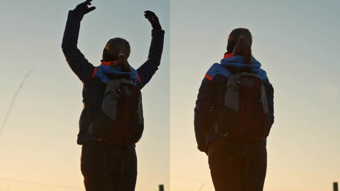 穿着保暖衣服的健康年轻女子的后视图，在乡村俯瞰日落的边境牧羊犬附近举起双臂进行调解