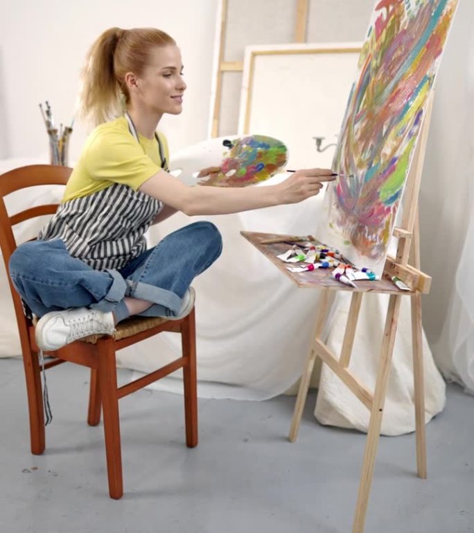 受启发的年轻女子绘制抽象图片，独自在工作室工作，垂直视频。快乐的女士坐在画布前，用鲜艳的色彩绘画，创