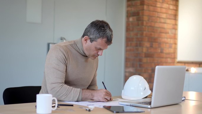 工程师建筑师用头盔、平板制作建筑图纸和蓝图。穿着衬衫，戴着眼镜，在办公桌上用铅笔创作房屋建筑平面图，