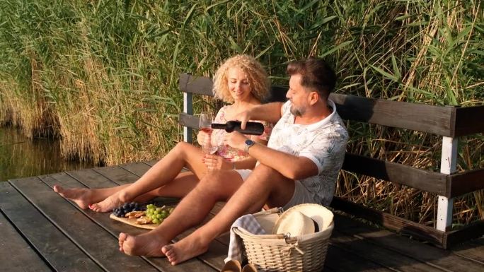 一对微笑的成年夫妇在日落时分坐在湖边的码头上喝酒。