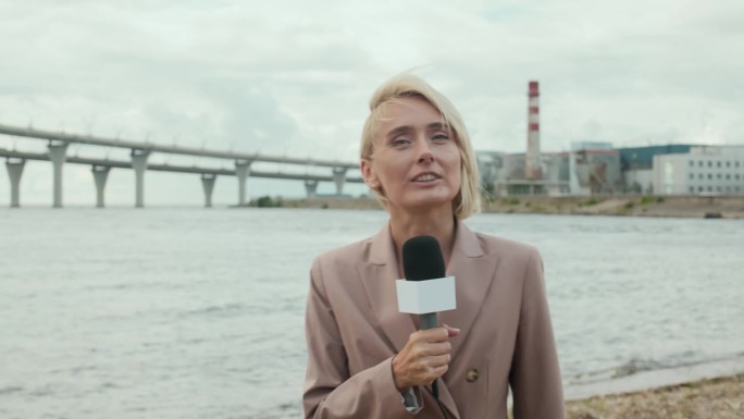 金发女子边做新闻报道边拿着麦克风走在水边