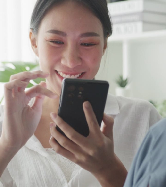 年轻的亚洲女性坐在家里客厅的沙发上玩智能手机。