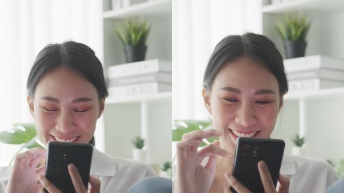 年轻的亚洲女性坐在家里客厅的沙发上玩智能手机。