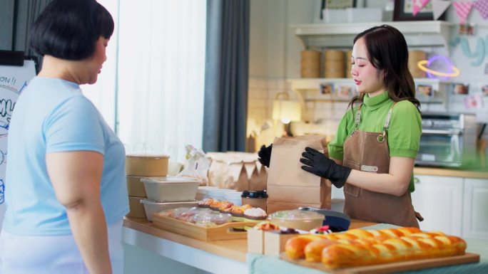 年轻的亚洲女顾客用手机为装满新鲜出炉的食品的纸袋付款。NFC二维码设备，payWave，非接触式支付