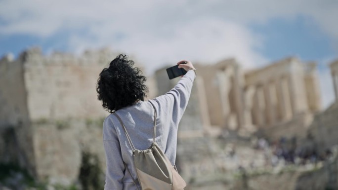 阳光明媚的一天，一名卷发女游客在阿略帕古斯山上用智能手机拍摄雅典卫城的照片