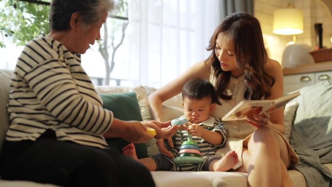 幸福的亚洲家庭生活方式妈妈，奶奶和孙子，男婴玩玩具，开心地笑，在沙发上快乐地在客厅的大窗户，阳光温暖