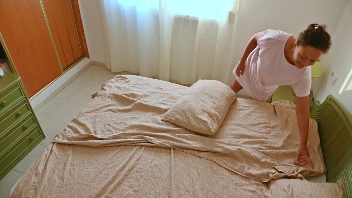 女人在明亮的卧室里铺床，在一个舒适的空间里展示日常生活和清洁，自然光从窗户透进来。