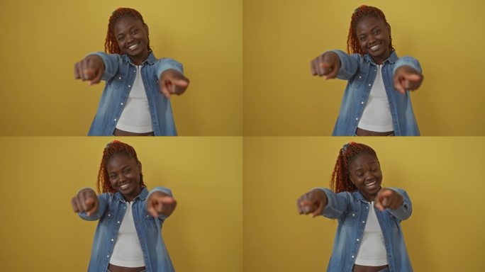 一个穿着牛仔衬衫的开朗的非洲裔美国女人，站在孤立的黄色背景上，微笑着指着你，用一个友好的手势向镜头示