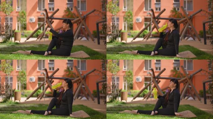 图为，一名女子坐在公寓楼院子外草地上的健身垫上，一边用黄色塑料瓶喝水，一边练习瑜伽。