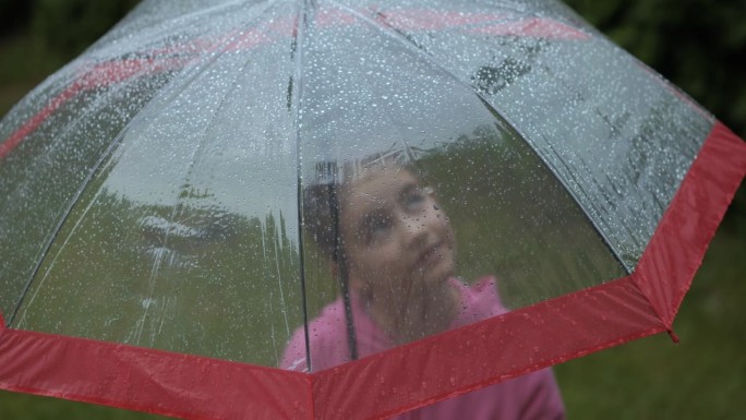 年轻可爱的女孩在雨中微笑，站在红色和透明的雨伞下。快乐的孩子仰望着雨中的天空。快乐的孩子在雨天户外享