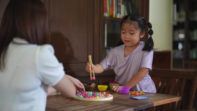 亚洲女性和一个孩子正在玩桌游。女性坐在桌子旁，面前有一碗食物。孩子站在她旁边，正在看桌游。