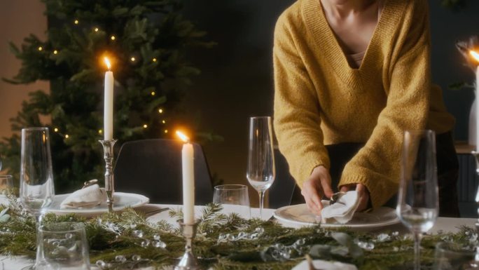 女人在圣诞节前将玻璃杯放在餐桌上，调整盘子上的餐巾。