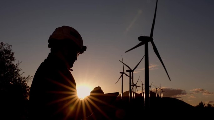 一名风力发电机维护技术员在风力发电机附近，专注地通过数字平板监控和控制系统，背景是日落时分。