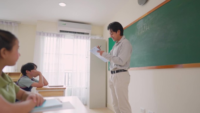 亚洲老师在小学教室里教学生。一群可爱的年轻孩子坐在桌子旁，学习并倾听他们的男老师讲解课堂内容。