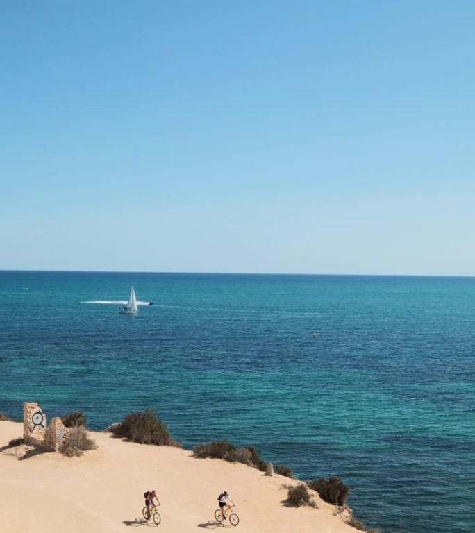 宁静的海岸场景，两名骑自行车的人沿着海边的沙滩小道骑行，俯视视角。普恩塔普里马，白色海岸，西班牙。