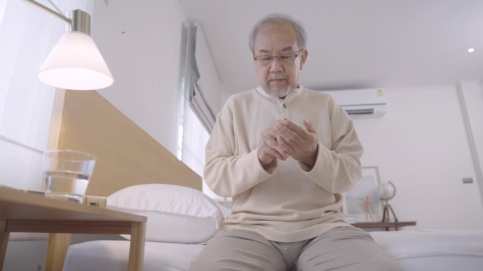亚洲老人坐在床上看书，然后在适当的时间吃药，然后继续在家看书
