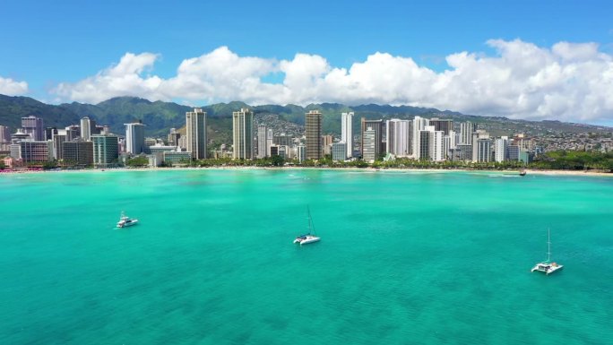 空中向下拍摄的白色帆船漂浮在蓝绿色的大海上，现代城市对多云的天空-瓦胡岛，夏威夷