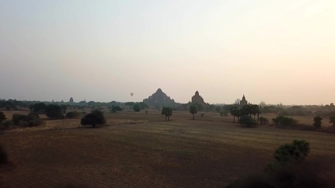 空中拍摄的树木在田野上的寺庙对天空在日出，无人机向前飞行的景观-蒲甘，缅甸