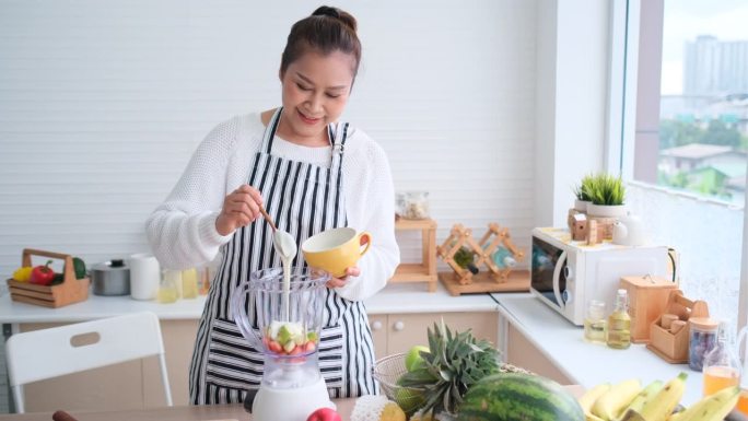 在厨房制作果汁的过程中，一位亚洲女士将酸奶放入含有混合水果的搅拌机中