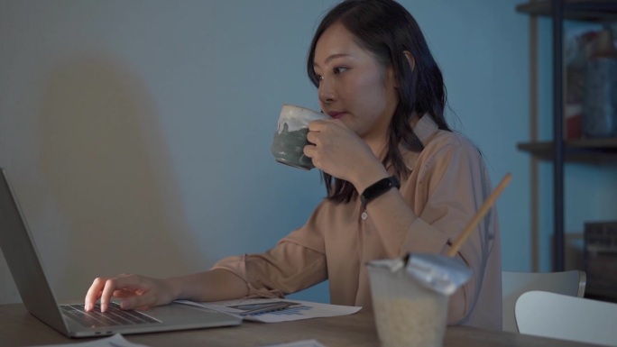 迷人的亚洲女人工作到很晚，喝咖啡与严肃的行动在桌子前的电脑显示器桌面在黑暗的工作场所，工作到很晚和努