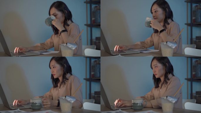 迷人的亚洲女人工作到很晚，喝咖啡与严肃的行动在桌子前的电脑显示器桌面在黑暗的工作场所，工作到很晚和努