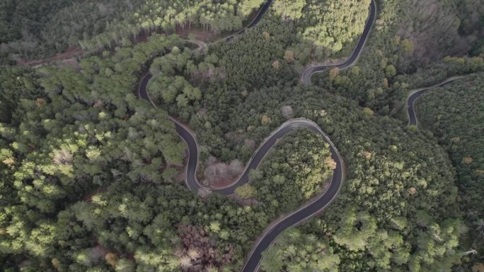 一架无人机飞越蜿蜒的山路。道路穿过陡峭的坡度，四周被高耸的山峰和郁郁葱葱的绿树环绕。广袤的山脉延伸至