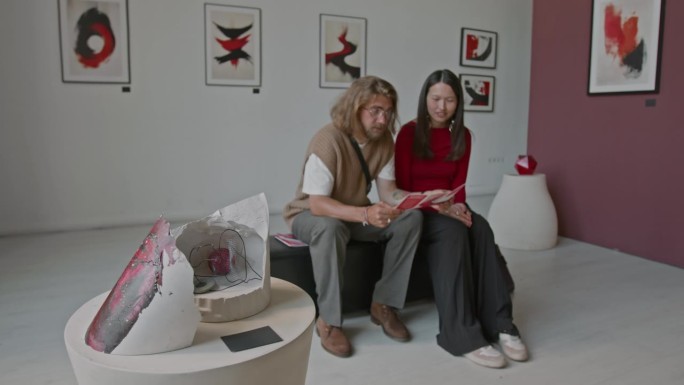 男人和女人坐在长椅上，阅读宣传册，欣赏画廊里的画作。