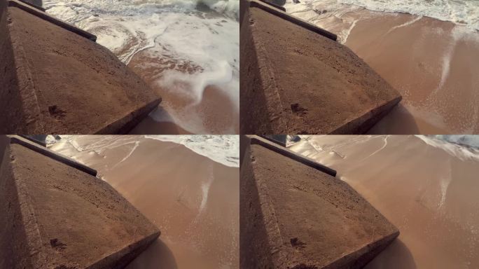 一段海滩上的混凝土人行道或障碍物，沙子与海水交汇的地方，突显了坚固结构与波浪流动之间的对比。