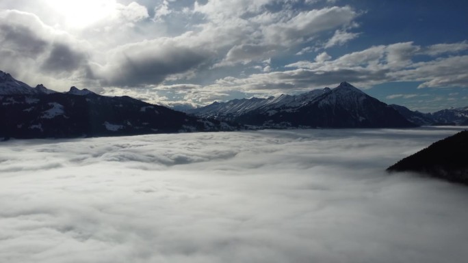无人机在阳光明媚的蓝天上飞越瑞士阿尔卑斯山的雪地，下面是一片云雾缭绕的海洋，点缀着几朵白云。