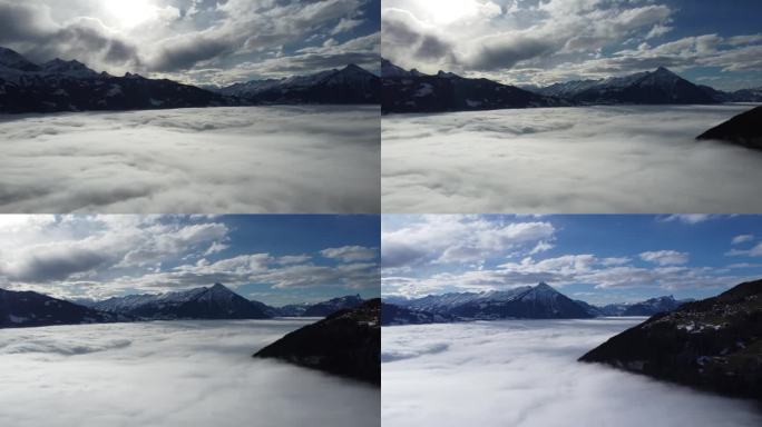 无人机在阳光明媚的蓝天上飞越瑞士阿尔卑斯山的雪地，下面是一片云雾缭绕的海洋，点缀着几朵白云。