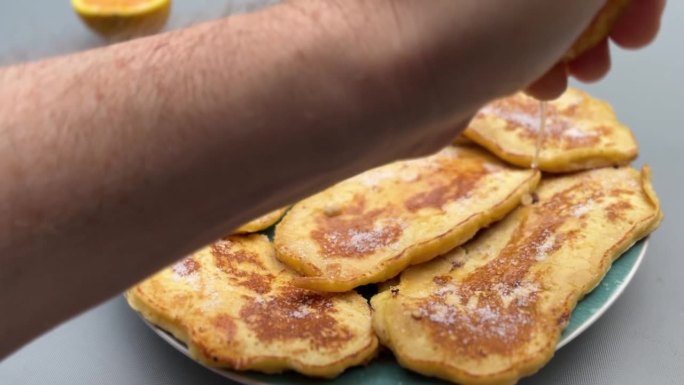 一只手在装满金色煎饼的盘子上挤柠檬，煎饼上轻轻撒上糖，并淋上新鲜橙汁。