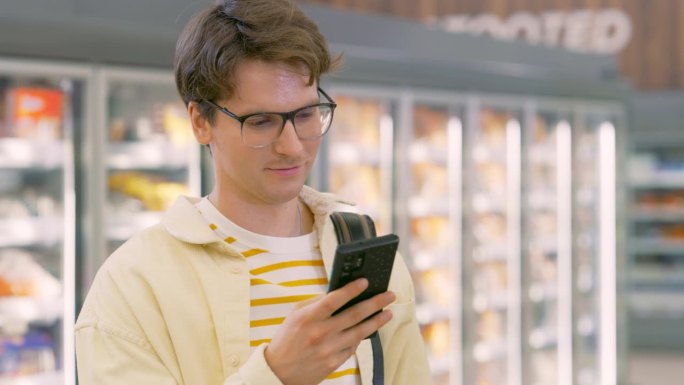 年轻帅气的男士在现代超市里悠闲地站着，手里拿着智能手机，使用手机仔细核对他还需要购买的早餐食品。