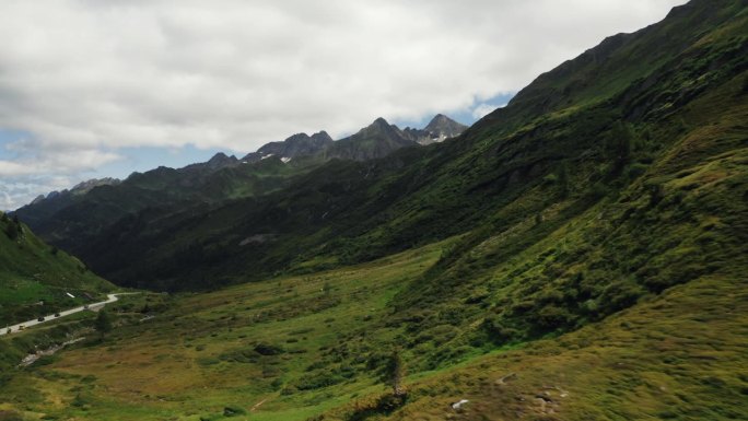 快速空中推进镜头 - 翠绿的阿尔卑斯山谷延伸在崎岖的山脊下，画面角落有蜿蜒的道路，天空多云。瑞士