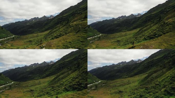 快速空中推进镜头 - 翠绿的阿尔卑斯山谷延伸在崎岖的山脊下，画面角落有蜿蜒的道路，天空多云。瑞士