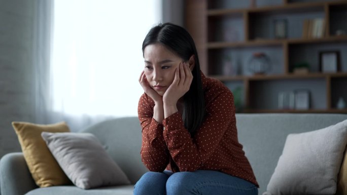 悲伤孤独的年轻亚洲女性正在家中的客厅沙发上坐着，感受着抑郁和心理上的痛苦。
