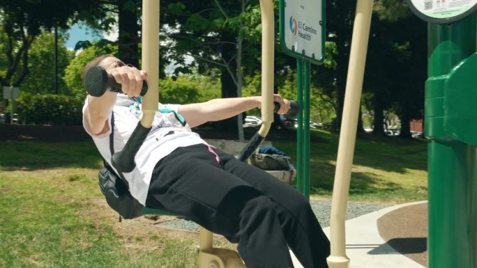 阳光明媚的夏天，老年女性在公共公园的户外健身器材上锻炼。