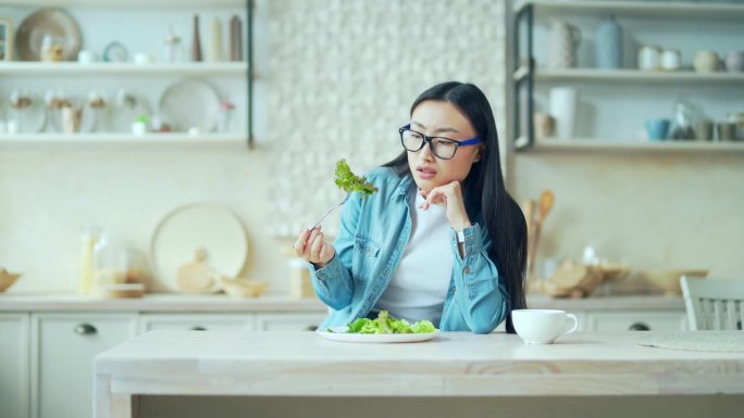 厌倦了节食减肥的年轻亚洲妇女坐在家里厨房的餐桌上，端着一盘沙拉