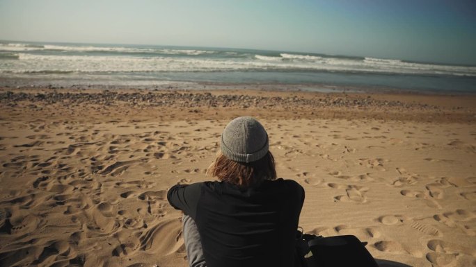 戴着针织帽的男子在阳光明媚的海滩上放松地欣赏着海景